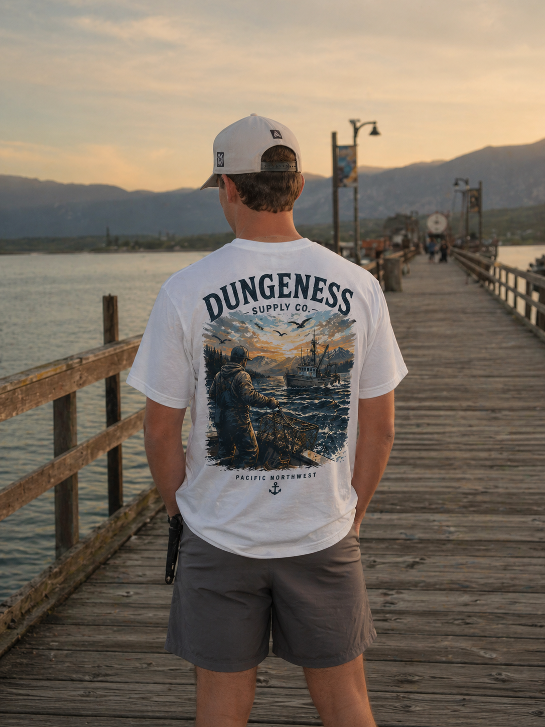White heritage tee with fisherman + mountains illustration, photographed at the pier at golden hour